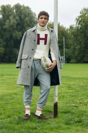Man in overcoat and sweatpants stands on grassy field holding rugby ball next to goal post wearing a sweater with the letter H.