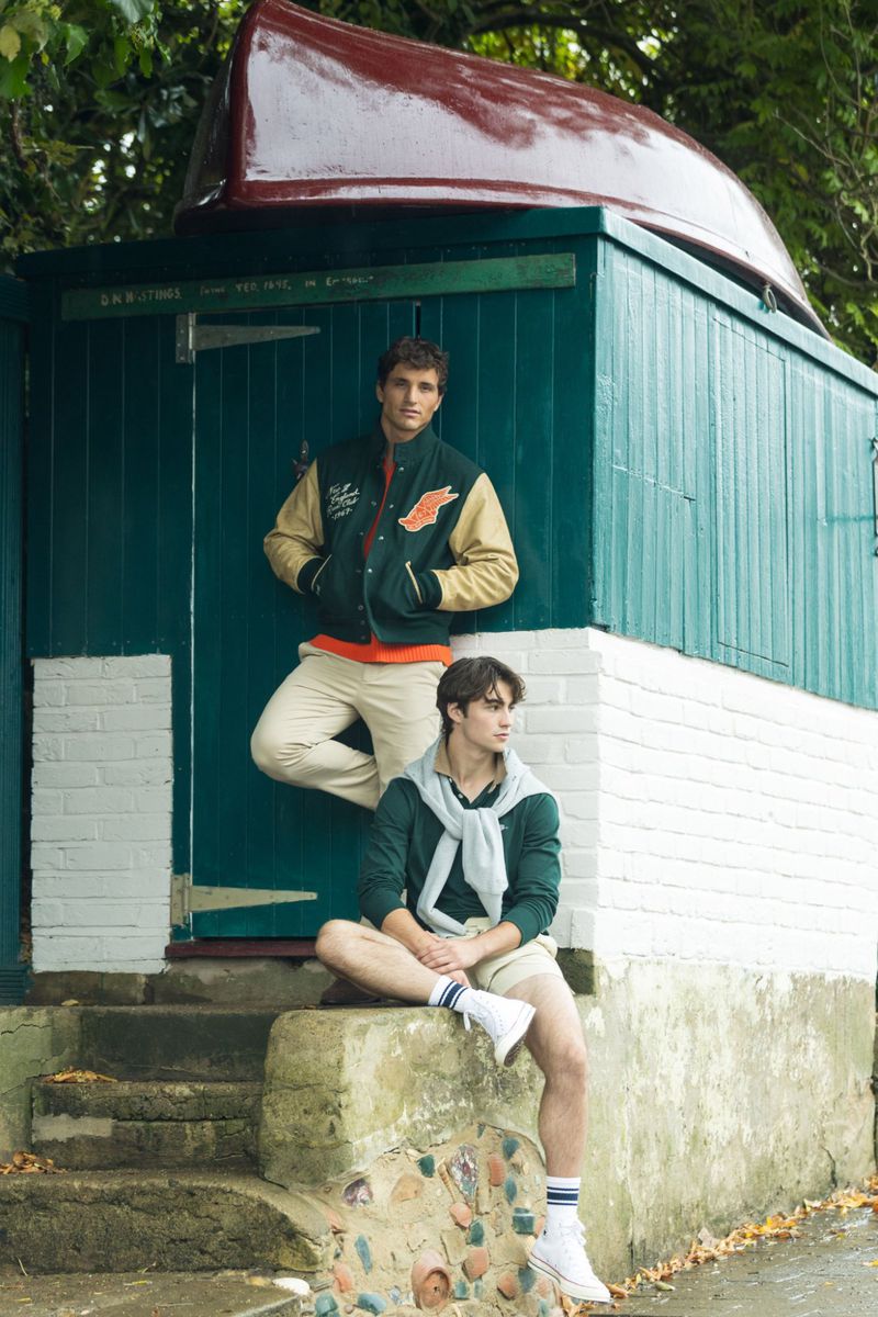 Two adult males pose outside a green boathouse; one leans against the boathouse, while the other sits on stone steps in neutral clothing.