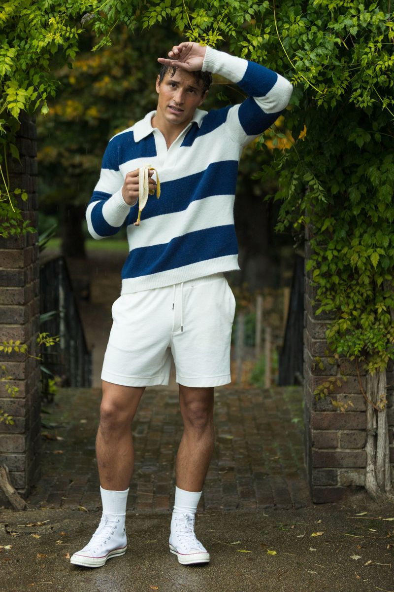 Man poses outdoors in casual wear, holding a banana, with a park-like setting and neutral tones, under a green leafy archway.