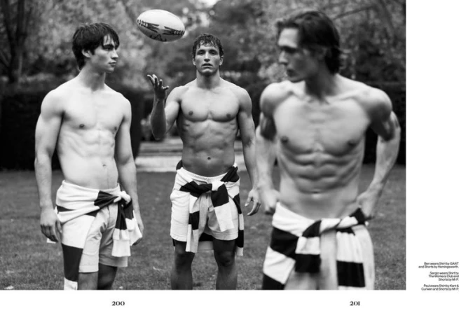 Three shirtless athletic men stand in a grassy field, one tossing a rugby ball in a black and white outdoor setting.