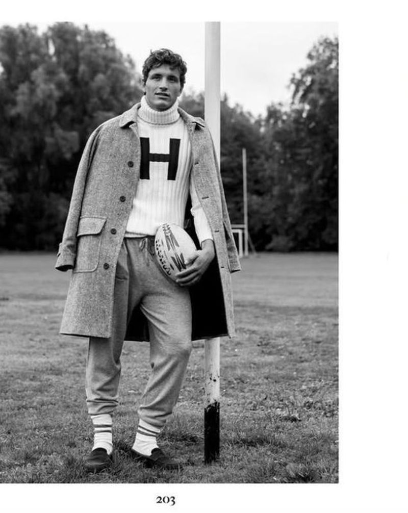 A man in a sweater with the letter H stands with a football in a grassy outdoor setting, wearing sweatpants and a jacket.