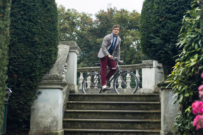 Adult male poses with a bicycle on stone steps in a park-like setting. He is dressed in maroon pants and a grey blazer.