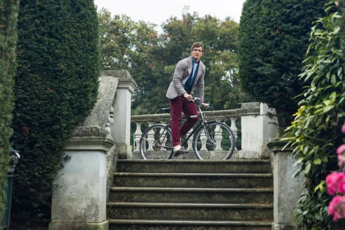 Adult male poses with a bicycle on stone steps in a park-like setting. He is dressed in maroon pants and a grey blazer.