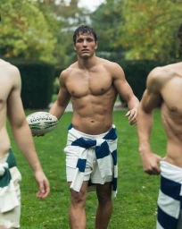 Shirtless man holding a rugby ball while walking on a grassy field with white shorts and a striped sweater tied around his waist.