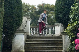 Man in grey blazer and red trousers riding a bicycle down stone outdoor steps flanked by manicured hedges for Take Ivy