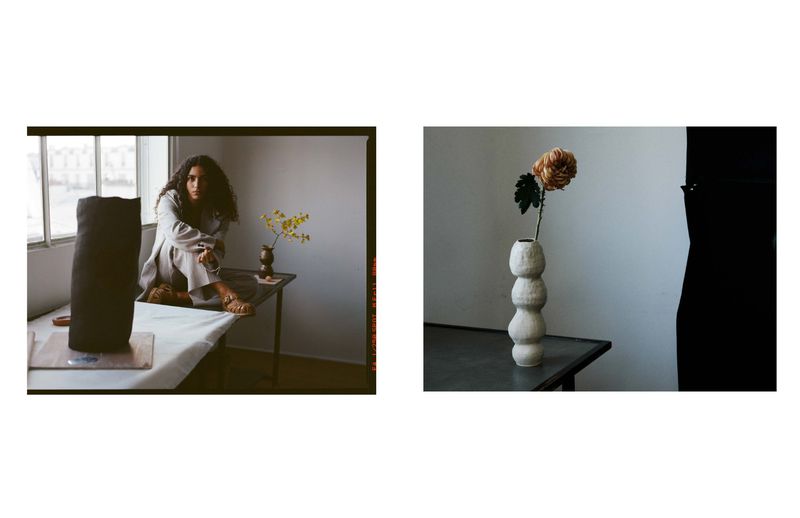 Indoor shot of a woman sitting on a table next to a sculpture and flowers. The setting includes a window and soft, natural lighting.