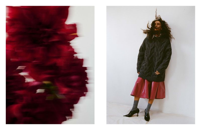 A woman with curly hair stands in front of a white wall wearing a sweater and skirt, with blurred red flowers in the left frame.