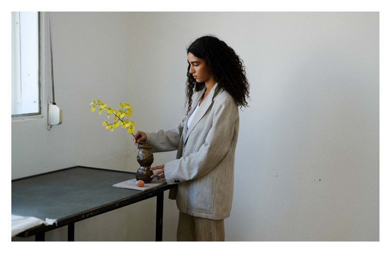 Woman arranging yellow orchid stems in a ceramic vase on a table in a bright room, wearing a neutral blazer and skirt.