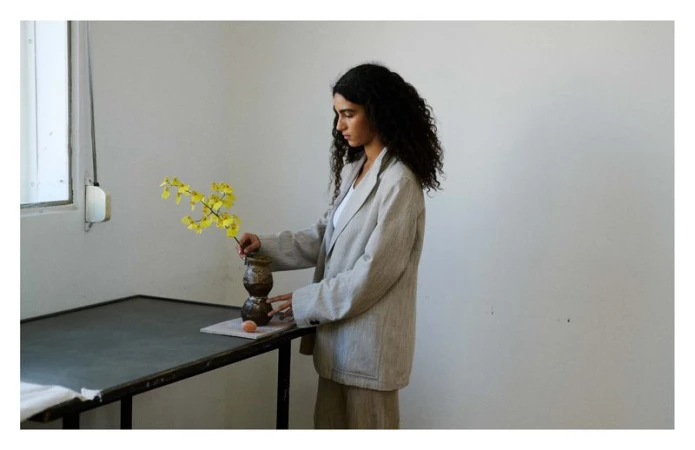 Woman arranging yellow orchid stems in a ceramic vase on a table in a bright room, wearing a neutral blazer and skirt.