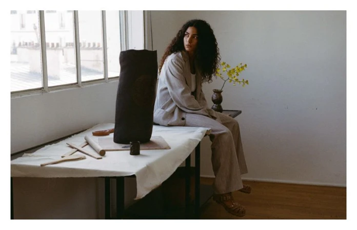 Adult female in a beige suit sits on a table next to a dark vase in a brightly lit room with a view of buildings outside a window.