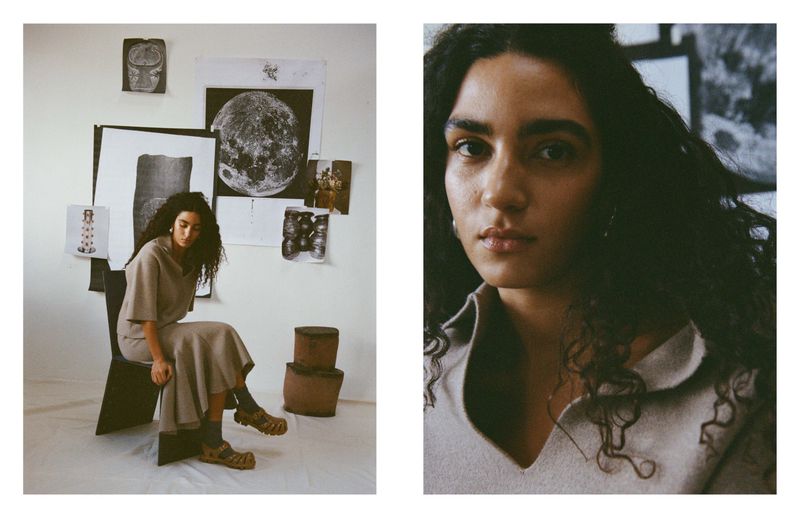 A woman with curly hair sits on a chair in neutral clothing, indoors, with various art pieces and prints on the wall behind her.