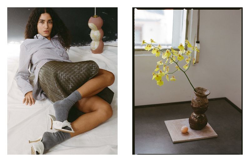 Fashion shot of a woman in a striped shirt and woven skirt reclining on a white sheet with a lamp, next to a floral arrangement.