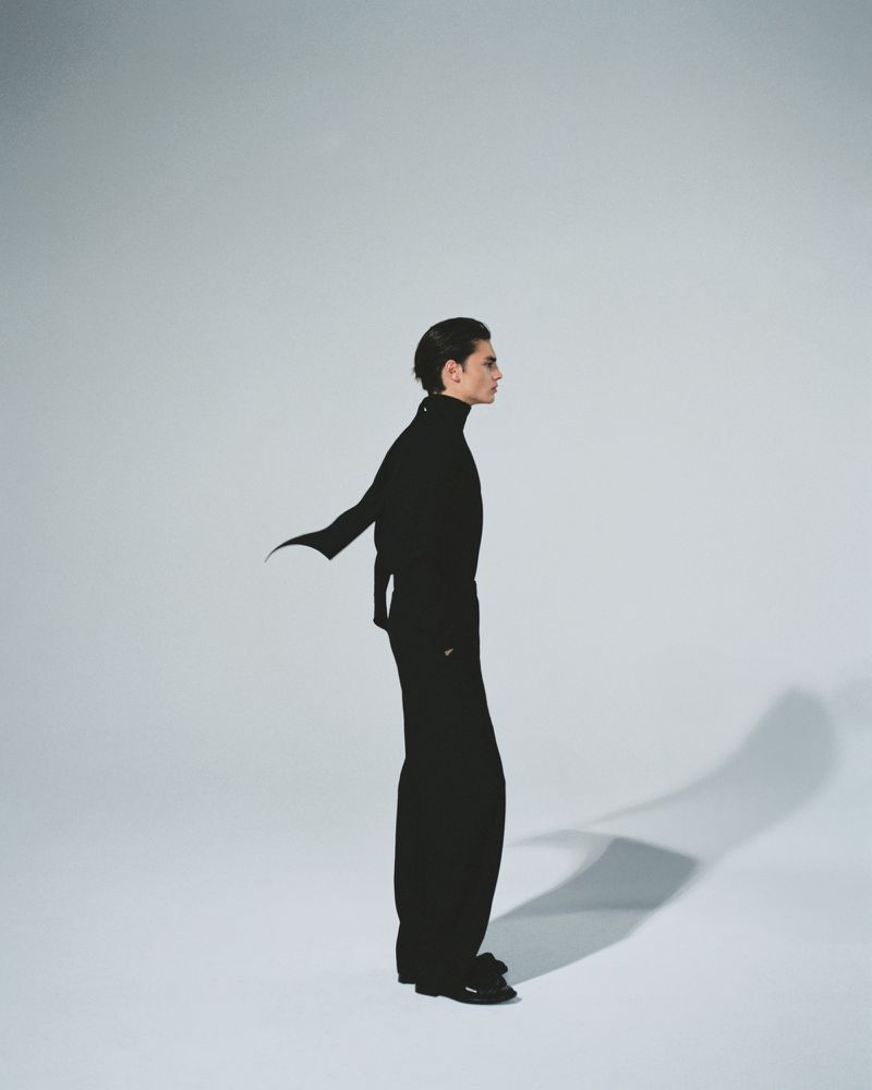 Side profile of a slender adult male in an all-black turtleneck outfit, posing in a photo studio against a solid light gray background.