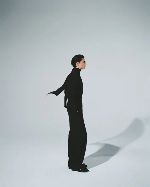 Side profile of a slender adult male in an all-black turtleneck outfit, posing in a photo studio against a solid light gray background.
