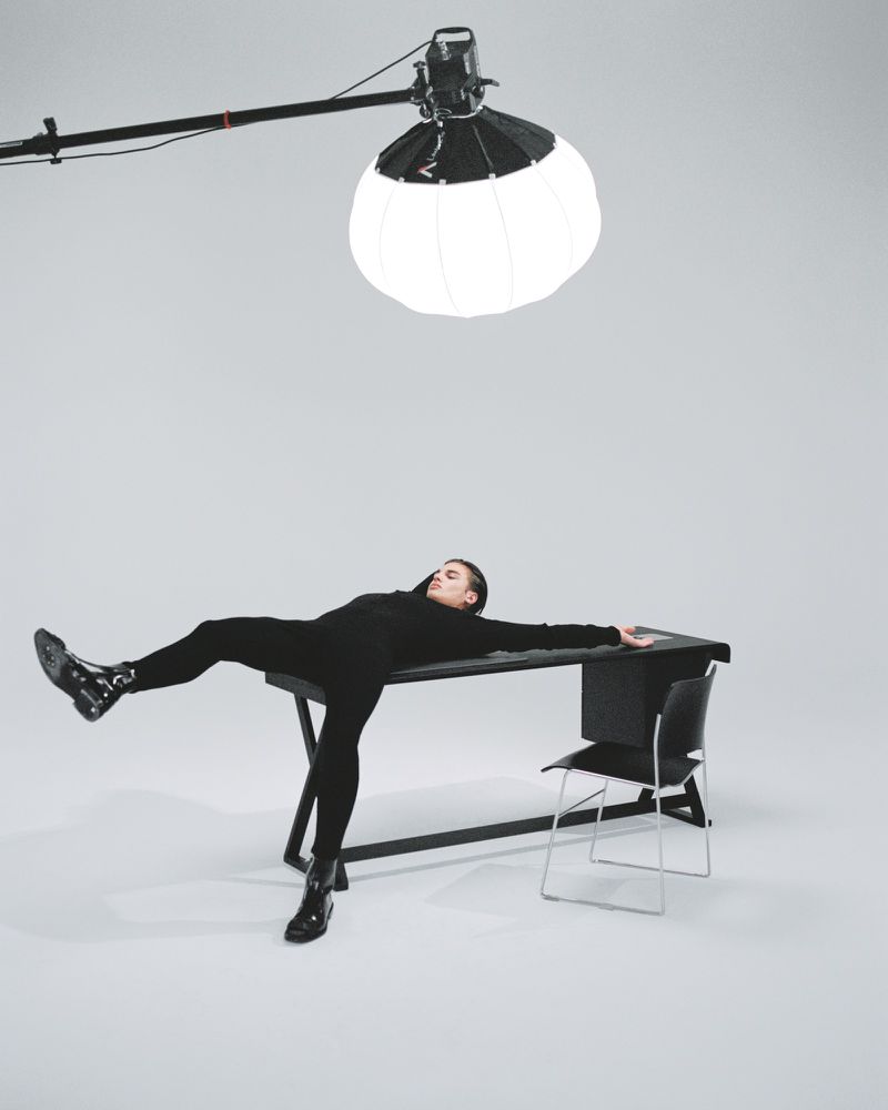 A model reclines on a desk in a studio setting, under soft lighting, wearing black clothing, with a chair and light stand in the background.