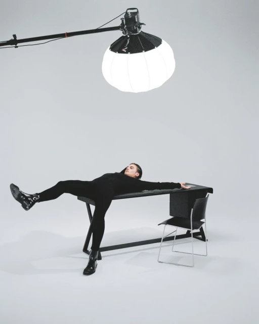 A model reclines on a desk in a studio setting, under soft lighting, wearing black clothing, with a chair and light stand in the background.