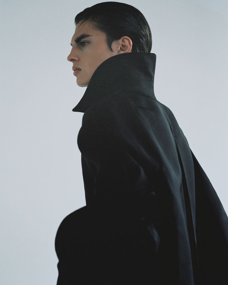 Adult male model in a black cape poses in a studio with a white backdrop. The image is cropped in portrait orientation, medium shot.