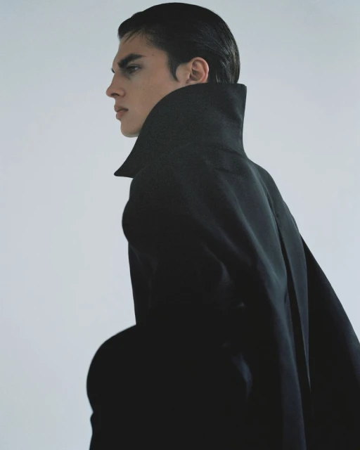 Adult male model in a black cape poses in a studio with a white backdrop. The image is cropped in portrait orientation, medium shot.