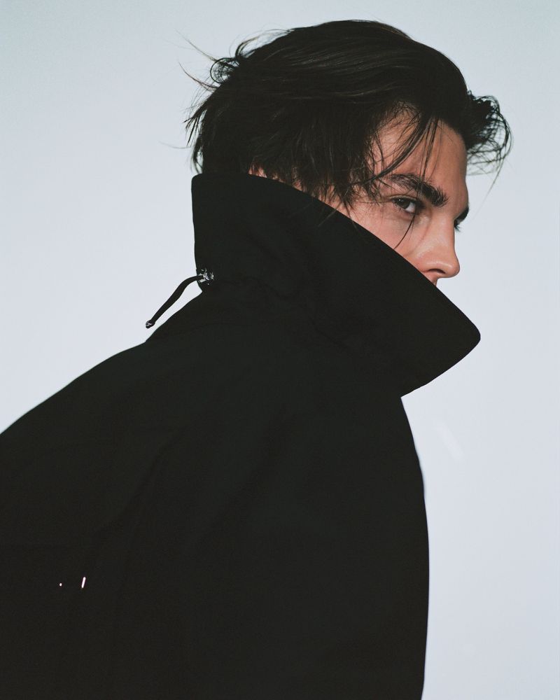 Portrait of a dark-haired adult male in a black coat with a high collar, looking to the side against a solid light background.