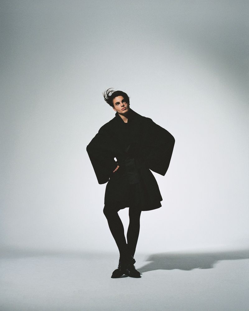Male model posing with hands on hips wearing an oversized black sculptural coat and black tights on a white studio seamless.