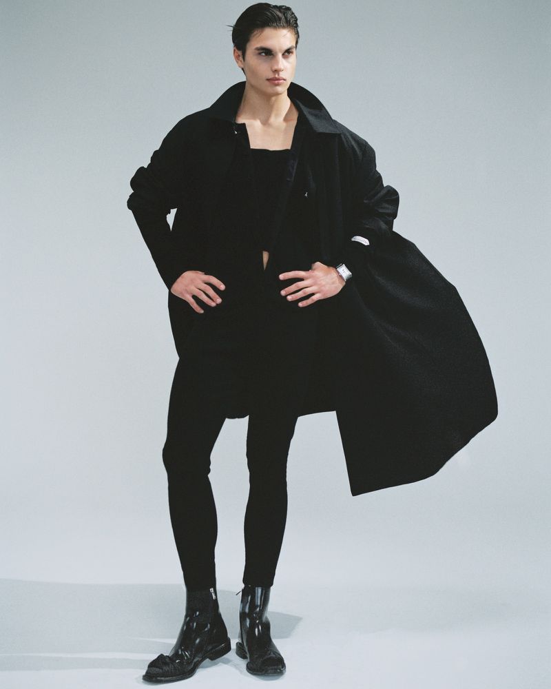Male model posing with hands on hips in an all-black outfit and flowing coat with glossy black boots against a light grey studio background.