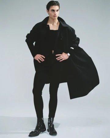Male model posing with hands on hips in an all-black outfit and flowing coat with glossy black boots against a light grey studio background.