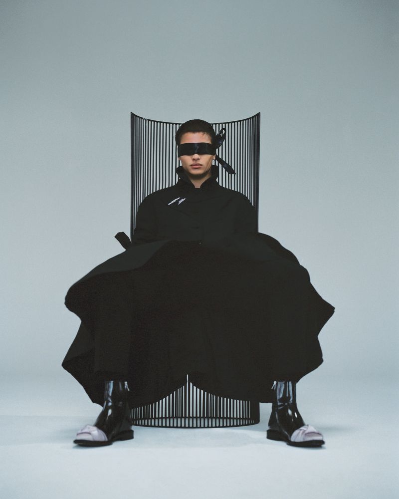 Male model in avant-garde fashion seated in sculptural chair, wearing a blindfold in a light studio setting, high fashion look.