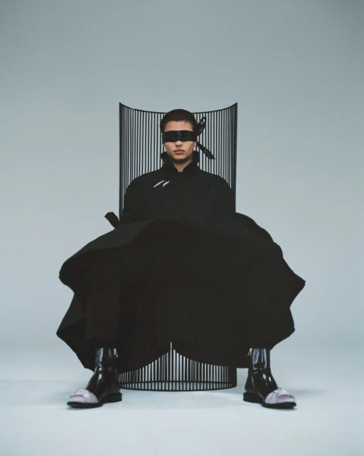 Male model in avant-garde fashion seated in sculptural chair, wearing a blindfold in a light studio setting, high fashion look.
