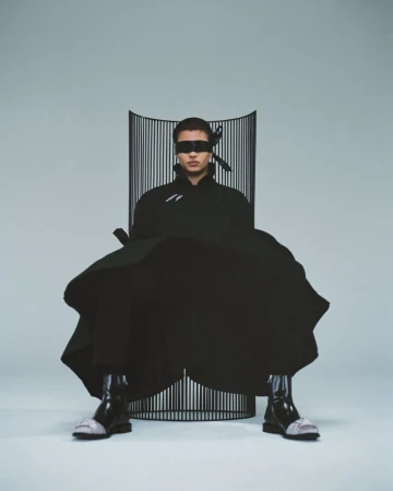 Male model in avant-garde fashion seated in sculptural chair, wearing a blindfold in a light studio setting, high fashion look.