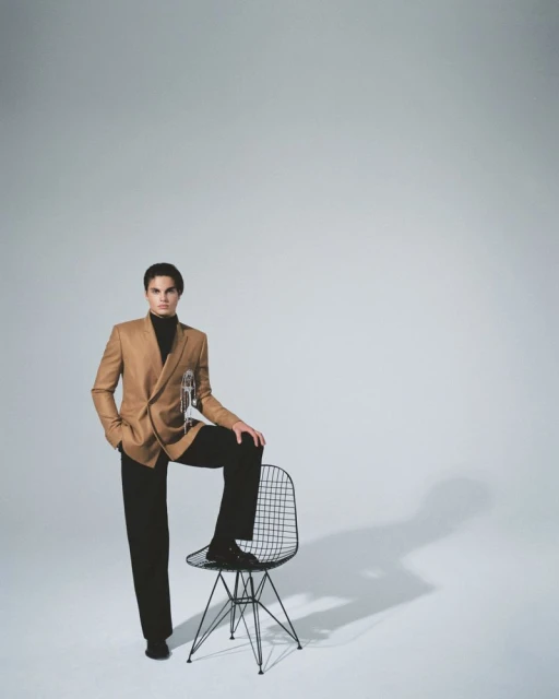 Full studio shot of a male model with tan blazer, black pants, and black turtleneck leaning on a black wire chair, fashionable pose.