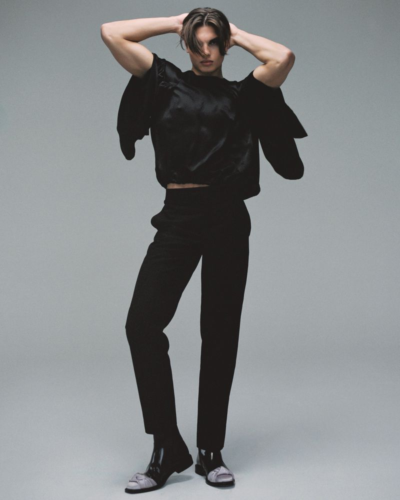 Model in black top and trousers with hands behind head against a grey studio backdrop