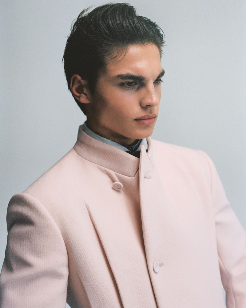 Portrait of a young adult male in a light pink coat, looking off to the side against a plain grey background. Fashion editorial.
