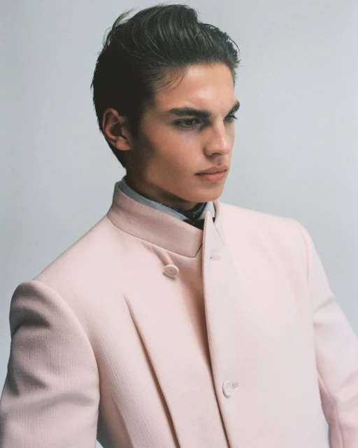 Portrait of a young adult male in a light pink coat, looking off to the side against a plain grey background. Fashion editorial.