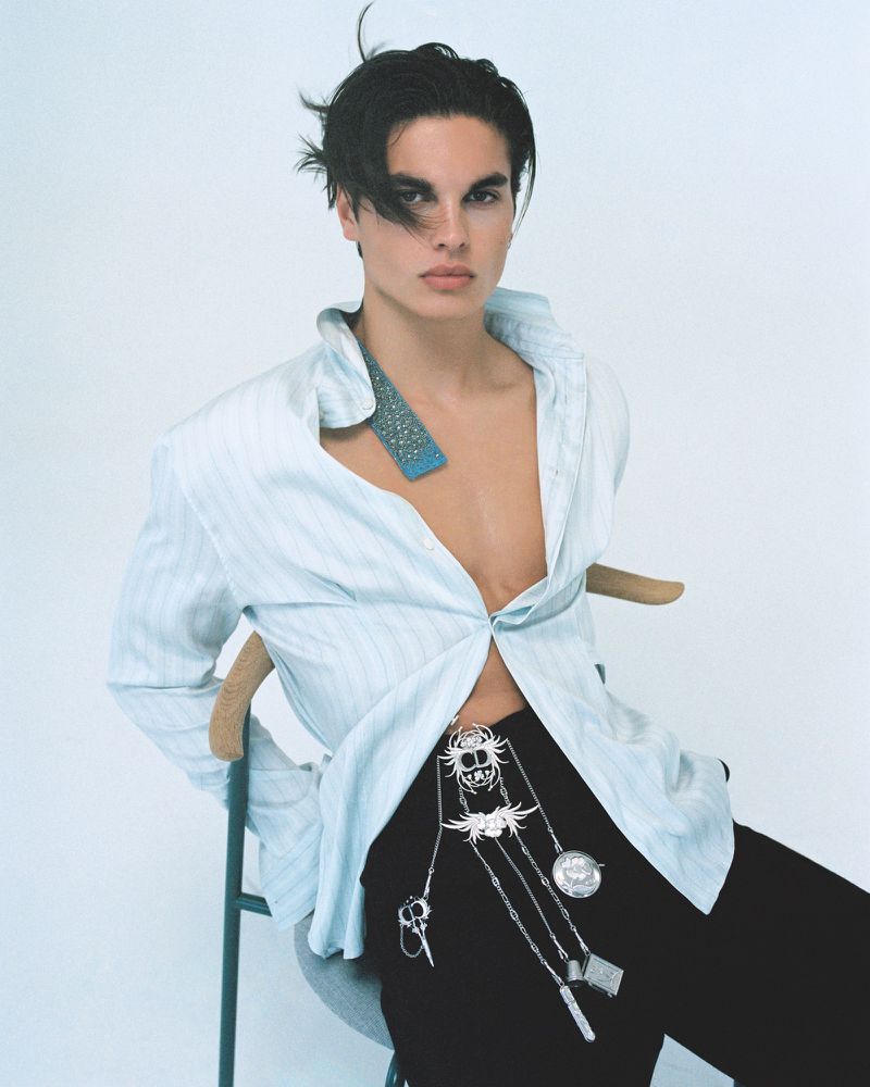 Portrait of a young adult male in a light blue shirt and black pants with jewelry, sitting on a chair in a bright white studio.