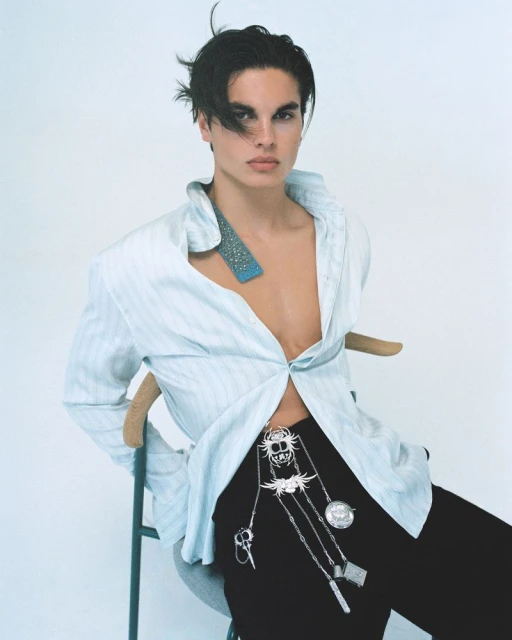 Portrait of a young adult male in a light blue shirt and black pants with jewelry, sitting on a chair in a bright white studio.