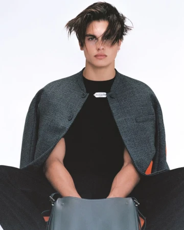 Male model with tousled dark hair seated against a white background wearing a draped grey jacket and black top with a leather bag.