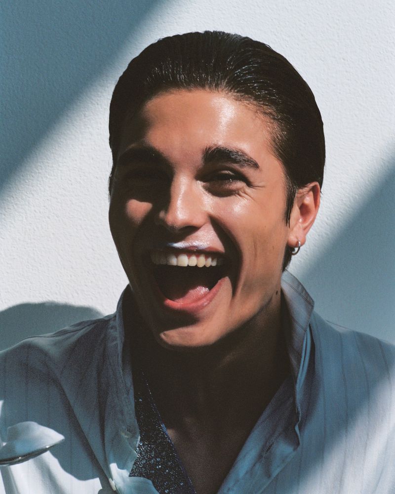 Close-up portrait of a laughing man wearing a light button-down shirt against a white wall with strong diagonal shadows.