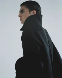 Profile portrait of a male model wearing a dark high-collared coat against a pale grey studio background.