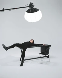 A man in an all-black outfit lying across a black desk under a large suspended lantern softbox in a minimalist studio.