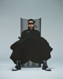 Model seated in a black wire chair wearing a black coat, shiny boots, and a black blindfold against a grey studio backdrop.