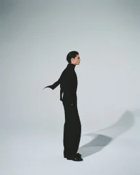Man wearing a black turtleneck and black trousers standing in profile against a plain white studio background.