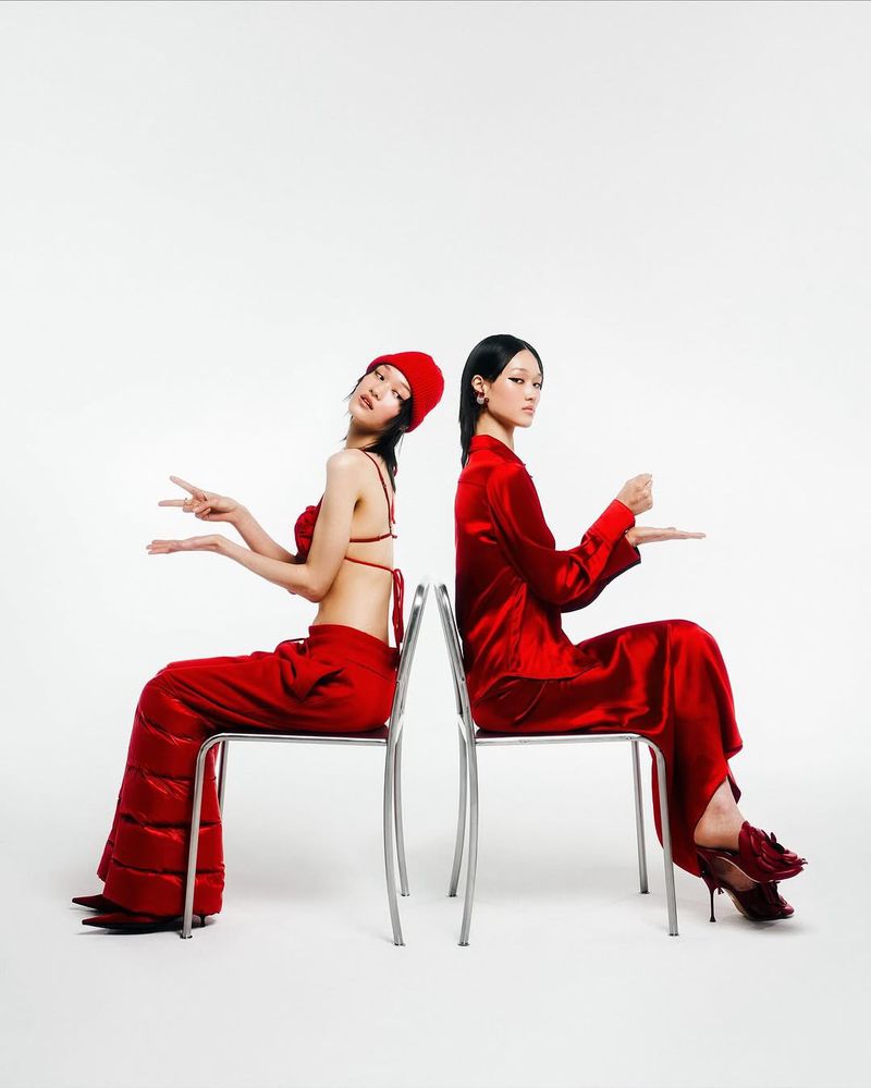 Two women in red outfits sit back-to-back on silver chairs in a studio setting. One wears a red beanie and the other has dark hair.