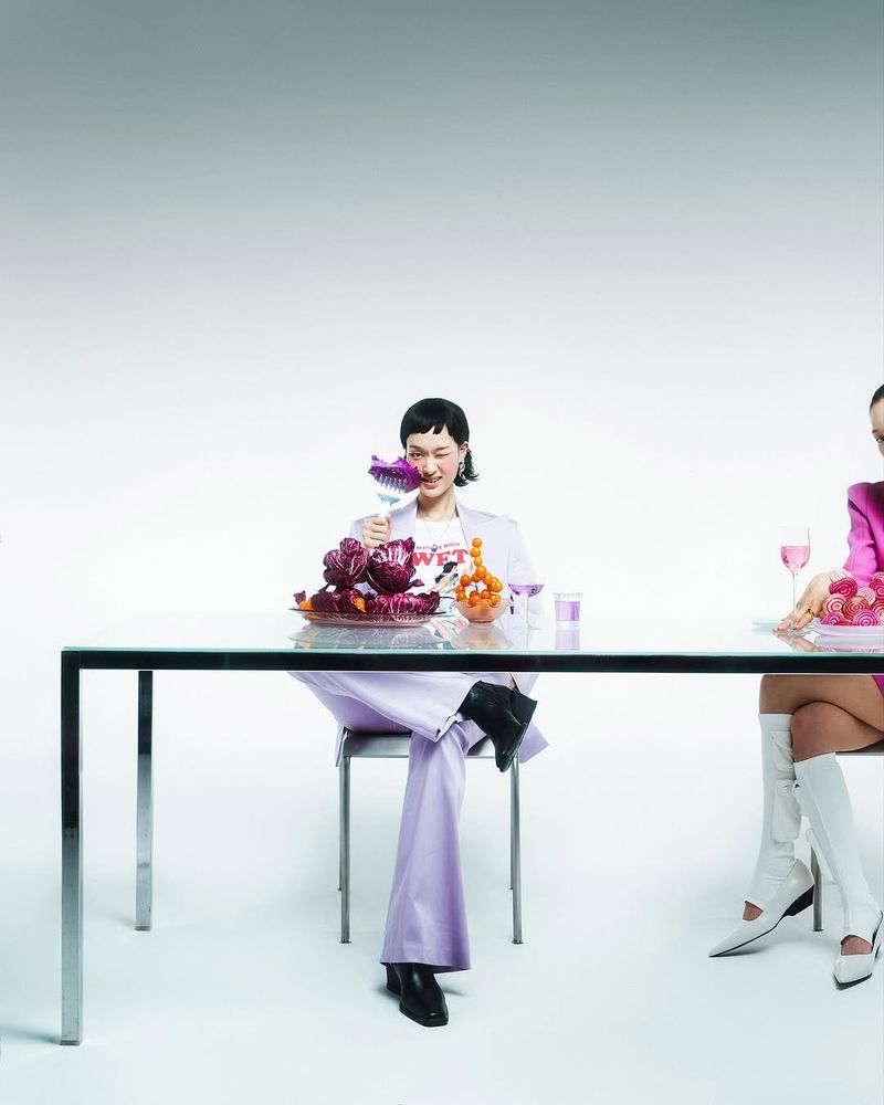 Two women at a table with purple cabbages and tangerines, wearing a purple suit and pink blazer with knee-high white boots in a studio.
