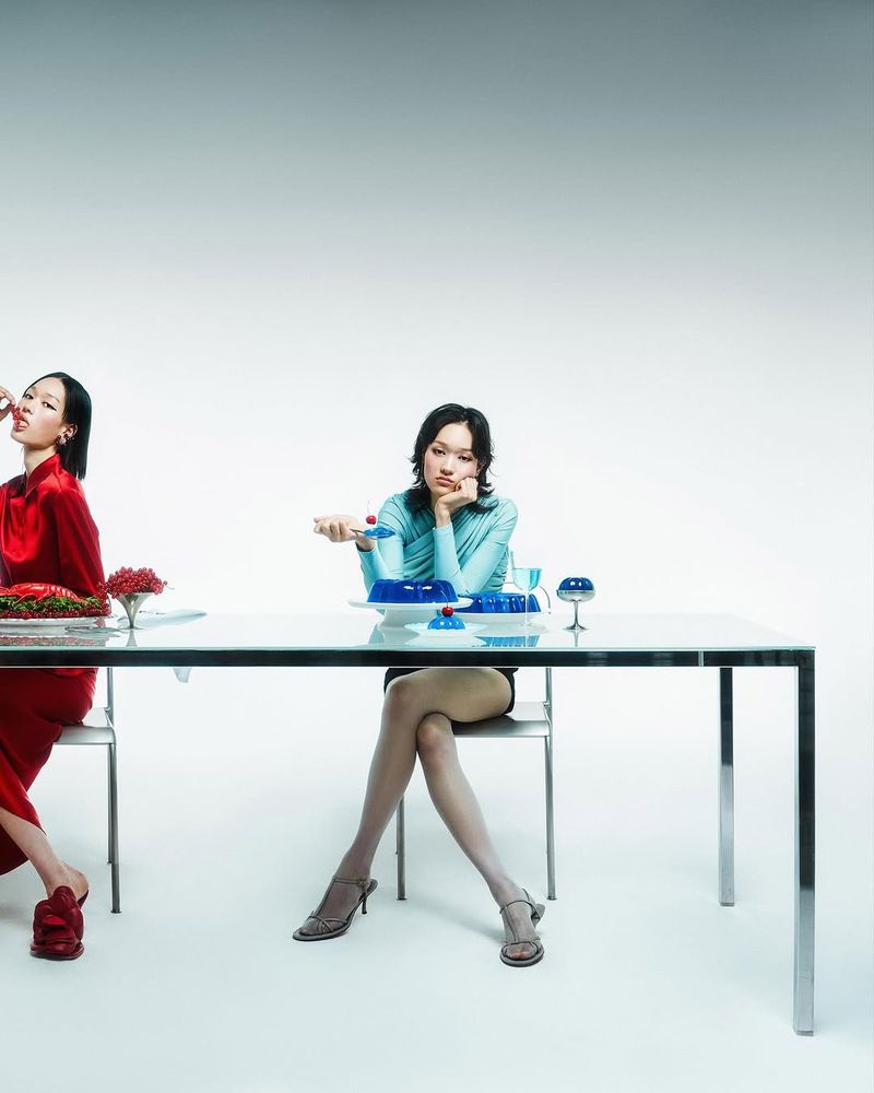 Two Asian women seated at table with blue jello dessert in brightly lit studio. One woman eats, the other rests her head on her hand.