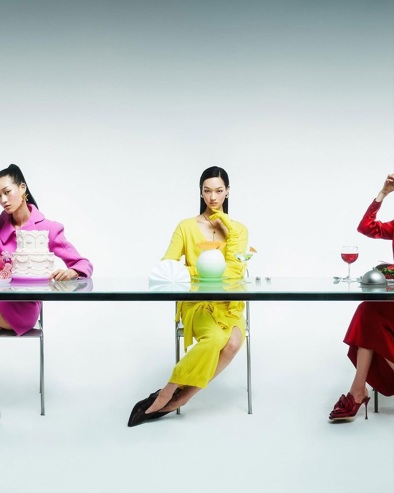 Three Asian women in colorful dresses sit at a glass table with a cake, fruit, and wine against a gradient white backdrop in a studio.