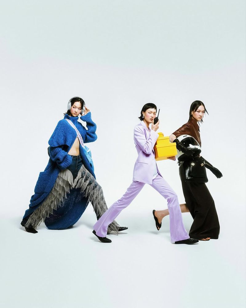Three women in fashionable, colorful outfits walk against a white backdrop, one holding a yellow box and another wearing headphones.