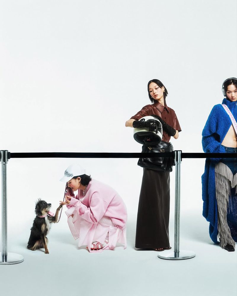Three stylish Asian women with an adorable dog, posing behind a black rope barrier in a studio setting, showcasing fashion apparel.