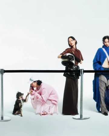 Three stylish Asian women with an adorable dog, posing behind a black rope barrier in a studio setting, showcasing fashion apparel.