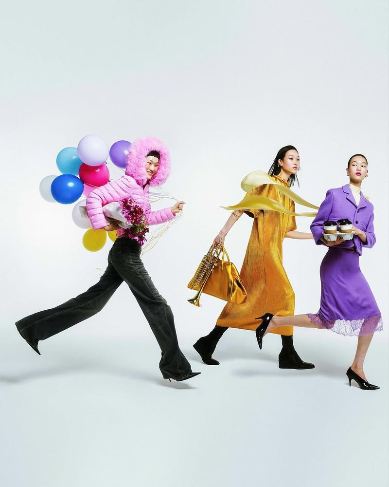 Three fashion models are running against a white backdrop with balloons, a trumpet, coffee cups, and flowers in a studio fashion shot.