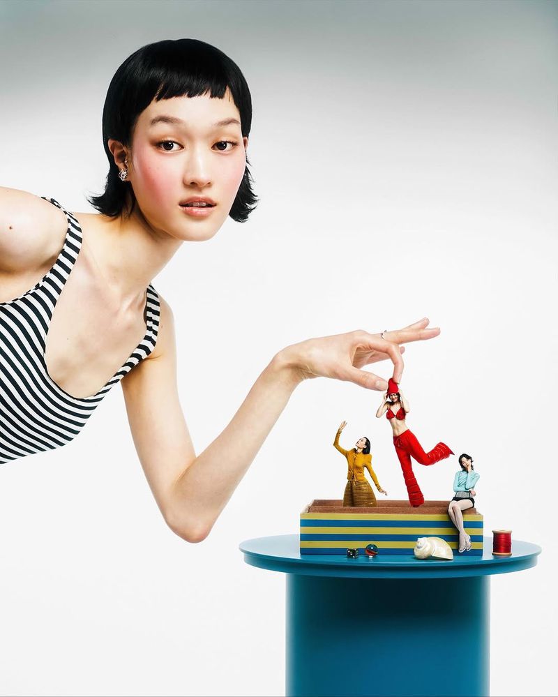 An Asian woman in a striped top interacts with miniature figures on a blue table, creating a surreal, fashion-forward studio portrait.